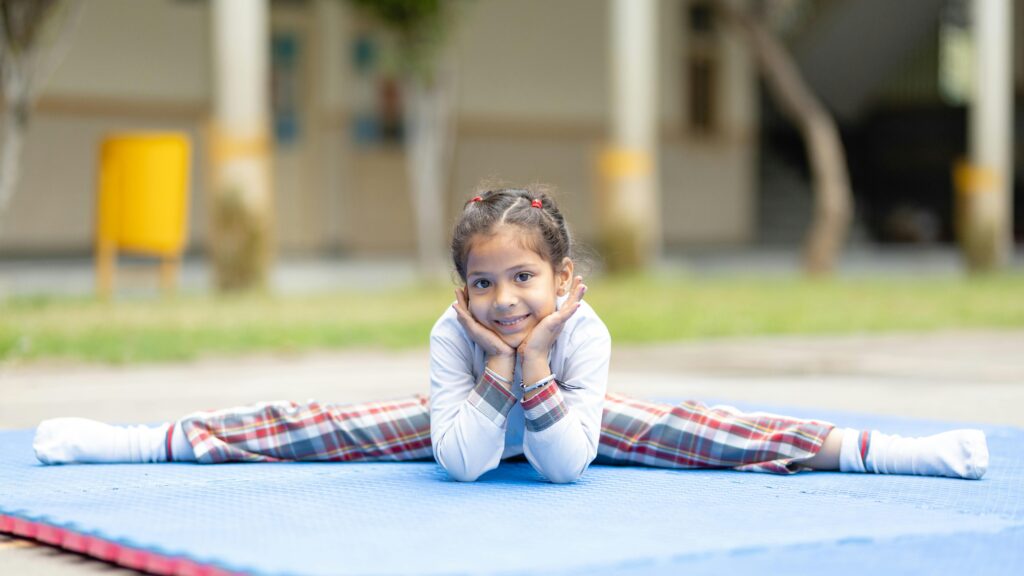 Young girl in school uniform performing a split on a blue mat outdoors, smiling.
