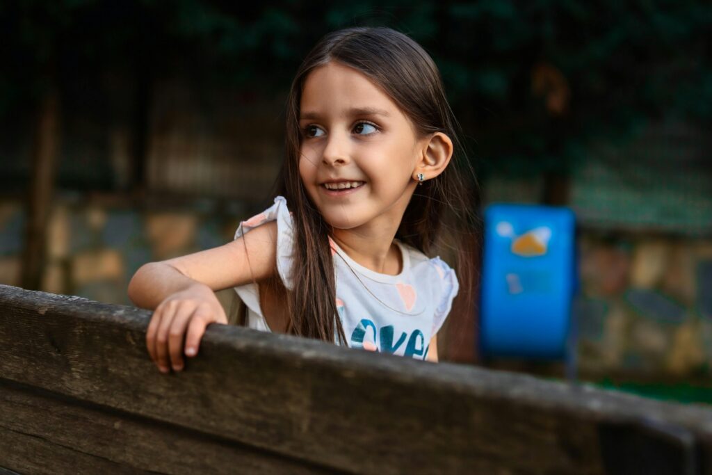 Young girl smiling while leaning on a wooden bench outdoors in a park setting.